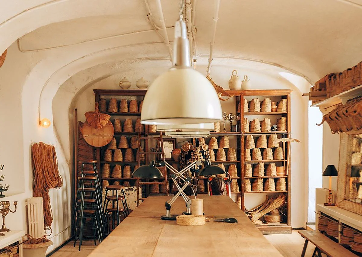 Imagen secundaria 1 - Interior del taller decorado con lámparas industriales. Encima, estantería antigua con cestos y bases de cabezas; y, a la derecha, jarrones de barro, y en la pared, cartel de una antigua taberna con más cabezas de animales, botijos y cestas. 